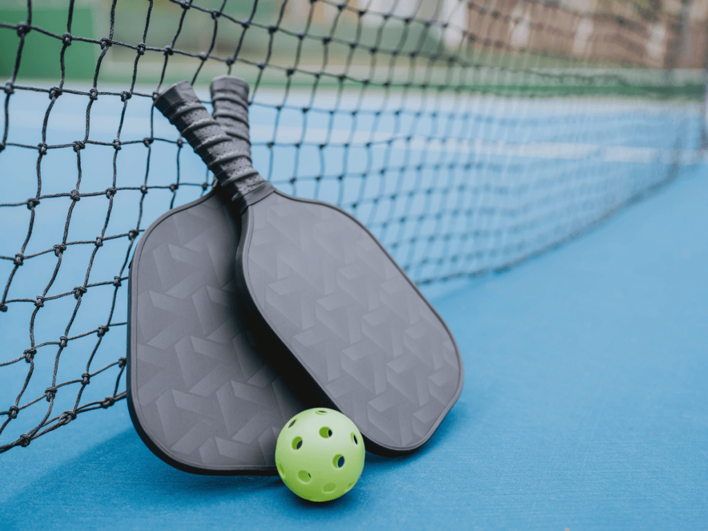 Best Beginner Paddles for New Pickleball Players 1 Two black beginner pickleball paddles and a green perforated ball rest against a net on a blue outdoor court, perfect for new pickleball players.