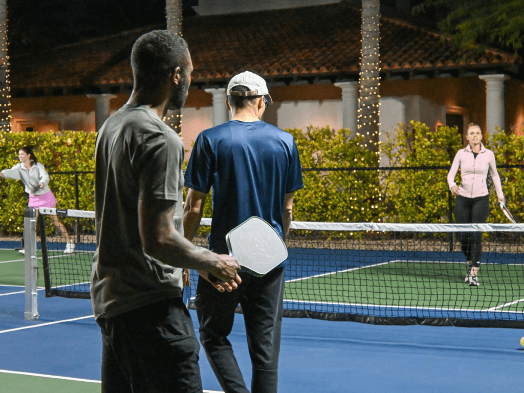 Best Beginner Paddles for New Pickleball Players 3 Four people, including new pickleball players, enjoy a nighttime game on an outdoor court as string lights twinkle around palm trees.