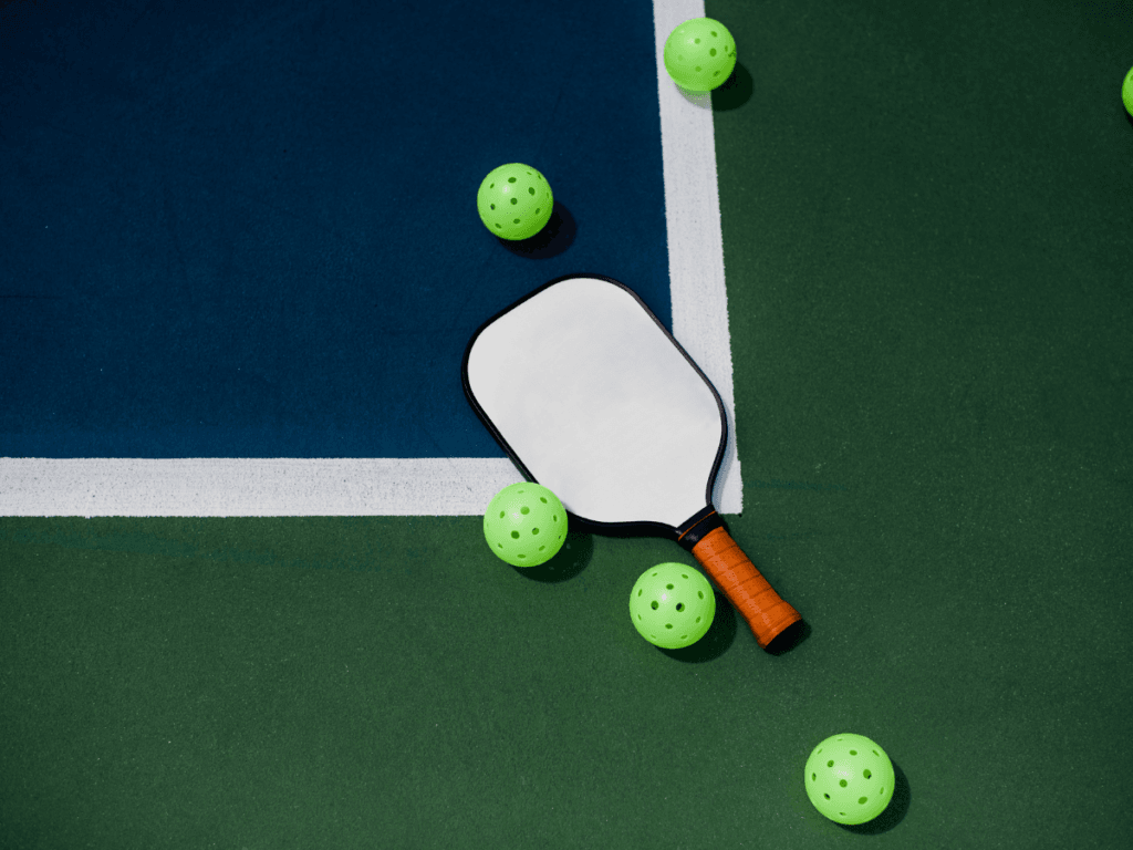 Best Beginner Paddles for New Pickleball Players 4 A pickleball paddle with a brown handle and five green pickleballs rests on a green and blue court near a white line—perfect for those seeking the best pickleball paddles for beginners.