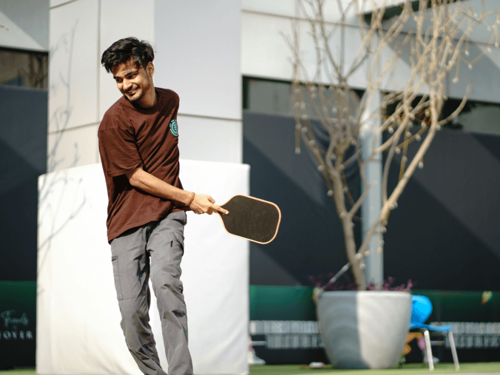 Best Beginner Paddles for New Pickleball Players 7 A man wearing a brown shirt and gray pants plays pickleball outdoors, smiling as he holds a beginner pickleball paddle—perfect for new pickleball players—with a large potted plant in the background.