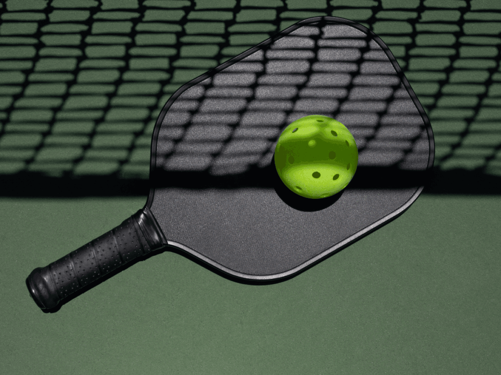 Best Beginner Paddles for New Pickleball Players 8 A black pickleball paddle and a green perforated pickleball lie on a green court surface, with the shadow of a net cast over them—an ideal scene for new players seeking beginner paddles to start their pickleball journey.