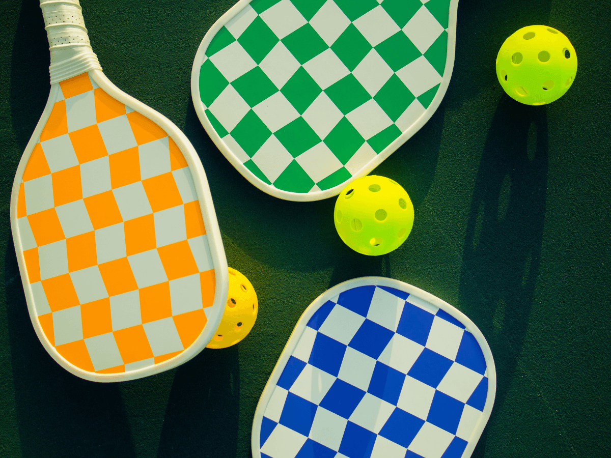 Three beginner paddles with checkered patterns in orange, green, and blue are arranged on a court with three yellow pickleballs, perfect for new pickleball players.