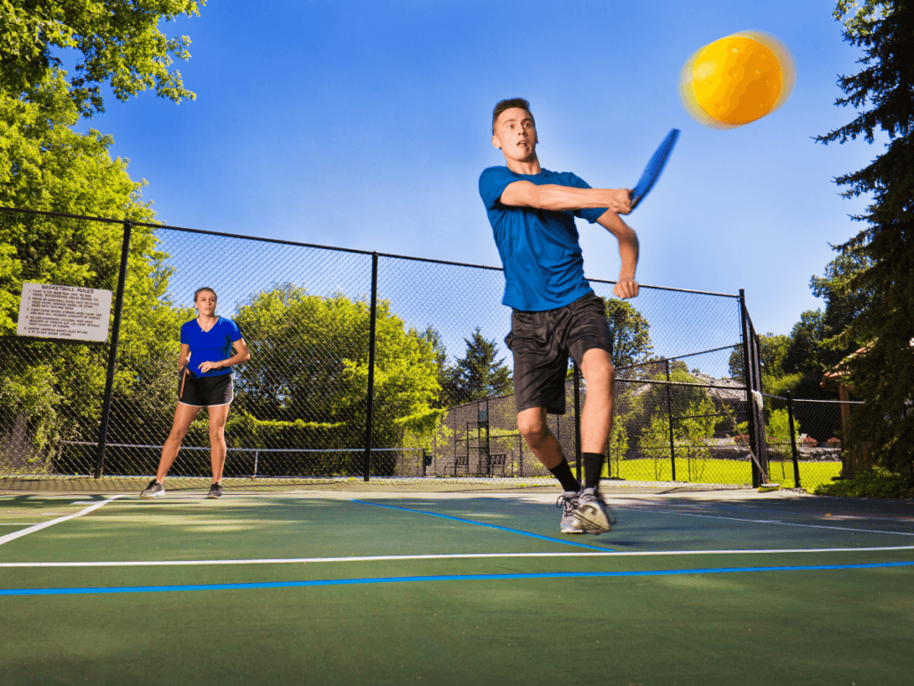 Top Dinking Drills That Improve Control and Consistency 2 A young man in blue demonstrates control as he hits a yellow pickleball with a paddle on an outdoor court, while a young woman stands ready behind him—showcasing consistency from their dinking drills.