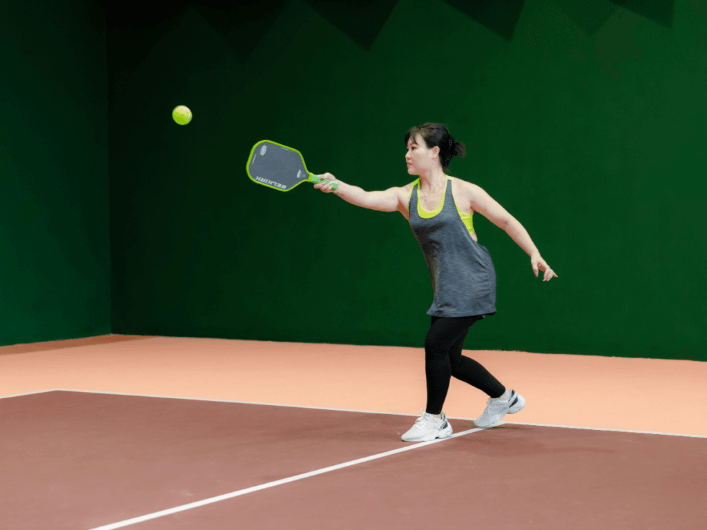 Top Dinking Drills That Improve Control and Consistency 3 A woman playing pickleball on an indoor court, reaching forward with a paddle to hit a ball in mid-air as she practices dinking drills to improve control and consistency in her game.