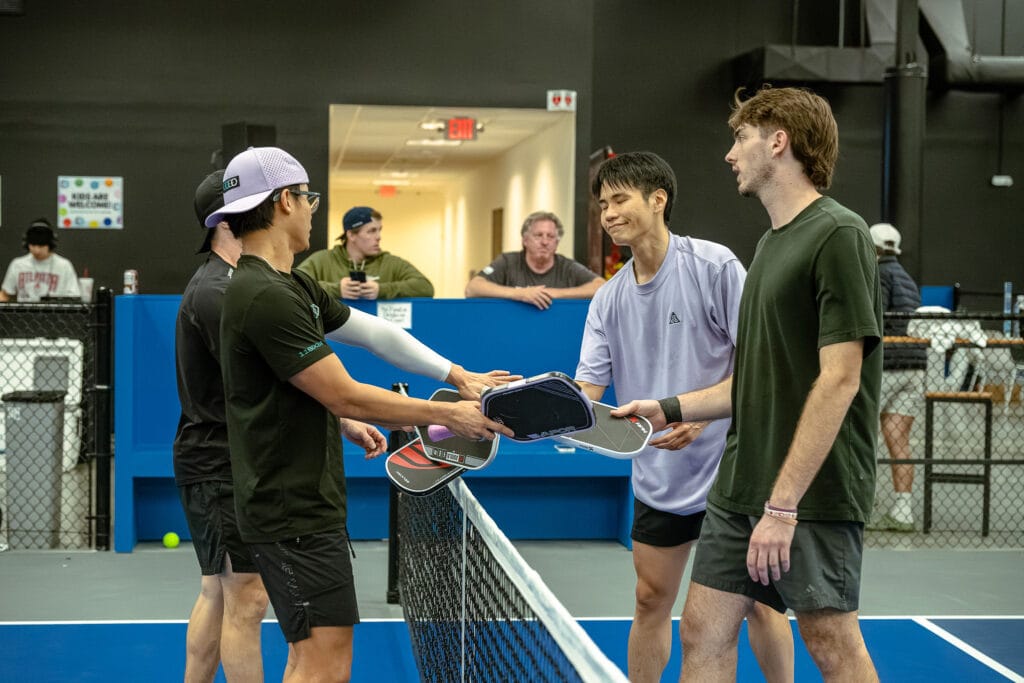 Four men on an indoor pickleball court tap paddles over the net at the end of a match, highlighting the camaraderie and sportsmanship fostered by Inclusive Programs, with people watching in the background.
