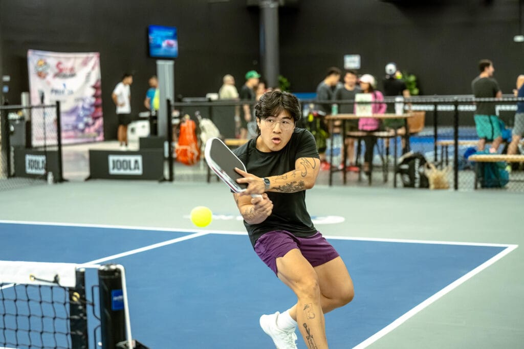 Tournaments 8 A person in athletic attire prepares to hit a pickleball with a paddle on an indoor court; other players and spectators are visible in the background.