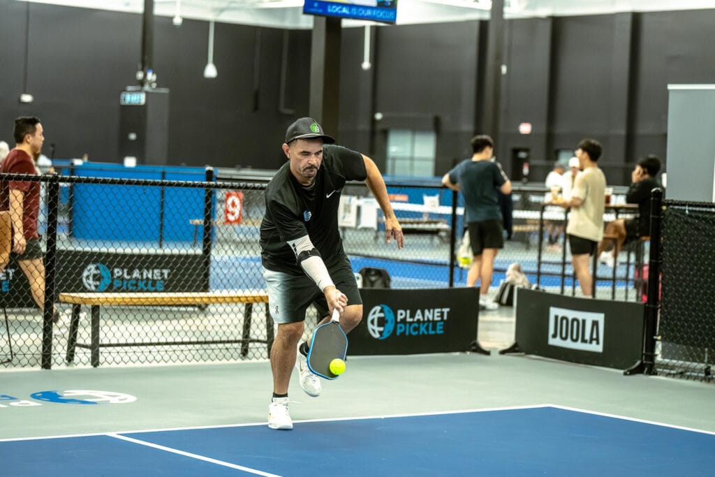 A man in athletic wear plays pickleball indoors, reaching forward with a paddle to hit a yellow ball on a blue court, with other players and benches in the background.