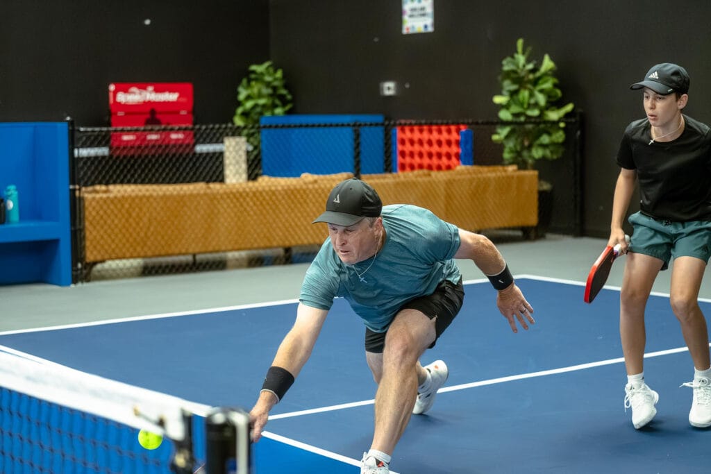 Partner Drills to Boost Your Pickleball Game: Effective Two-Player Exercises for Doubles Success 1 Two people playing pickleball indoors; the man lunges forward to hit a low ball near the net while the woman stands ready behind him.