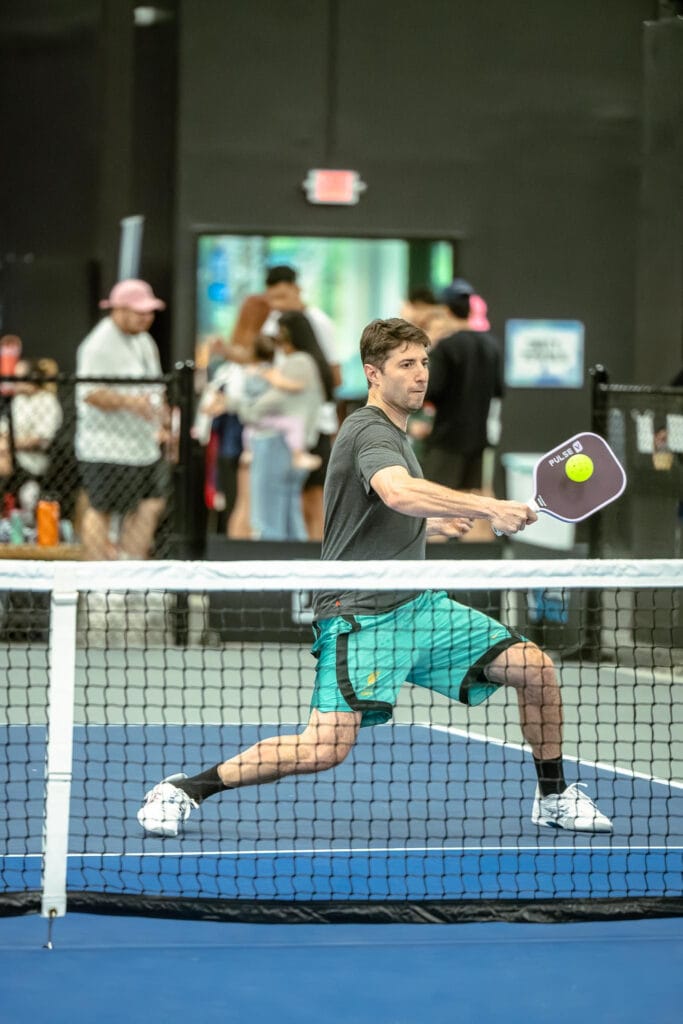 Tournaments 7 A man in athletic wear plays pickleball indoors, preparing to hit a ball with his paddle near the net; spectators are visible in the background.
