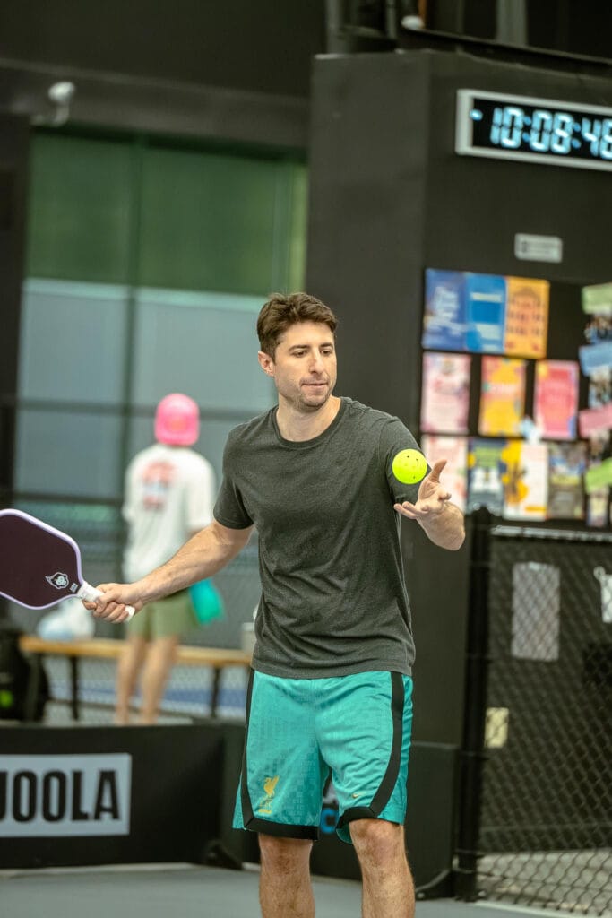 A man in a gray t-shirt and teal shorts prepares to serve a pickleball indoors, holding a paddle in one hand and tossing the ball with the other.