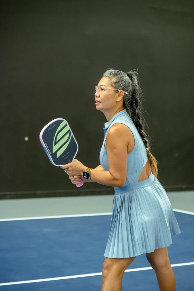 A woman in a blue dress stands on a pickleball court, holding a paddle and preparing to play.