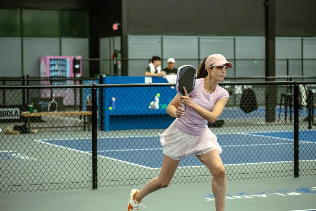 A woman in athletic wear plays pickleball indoors, holding a paddle and preparing to hit the ball near a fenced court.