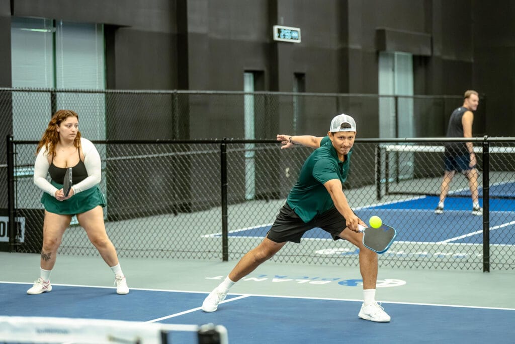 Partner Drills to Boost Your Pickleball Game: Effective Two-Player Exercises for Doubles Success 4 Two people play doubles pickleball indoors, one hitting the ball while the other stands ready; another game is visible in the background. This lively setting is perfect for private events.