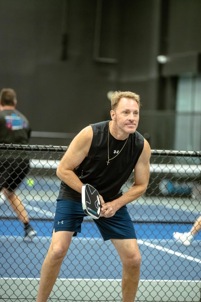 A man in athletic wear prepares to play pickleball on an indoor court, holding a paddle and standing near the net. Other players are visible in the background.