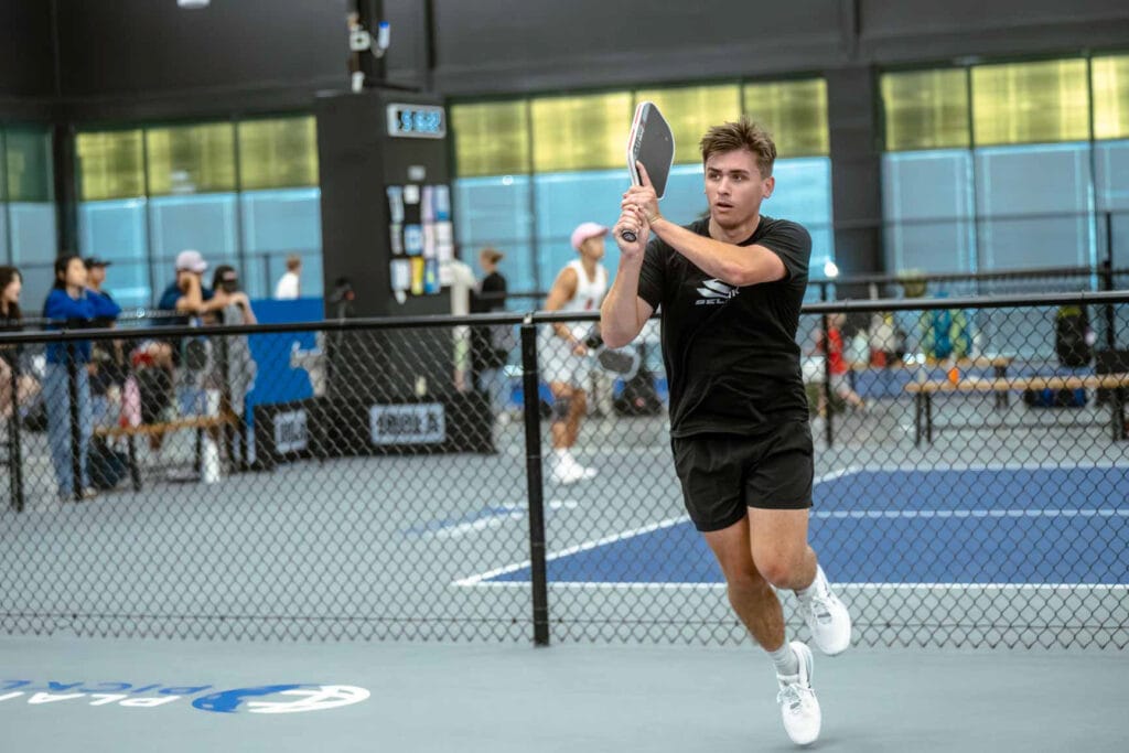 A young man in athletic attire plays pickleball indoors, preparing to hit the ball with a paddle near a netted court. Spectators are visible in the background.