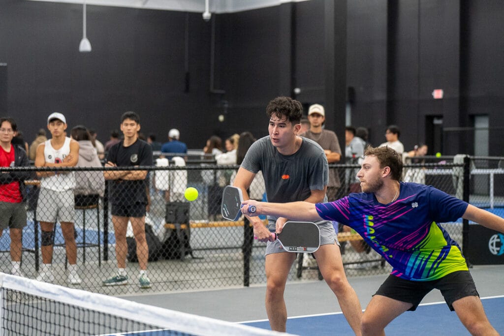 Two men playing pickleball indoors, one reaching to hit the ball with a paddle while spectators watch behind a black fence.