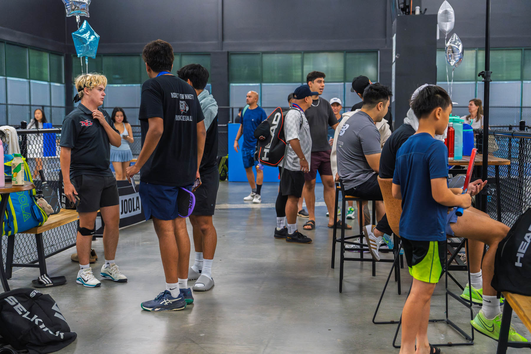 A group of people, including adults and children, gather inside an indoor sports facility with tables, benches, and sports equipment visible.
