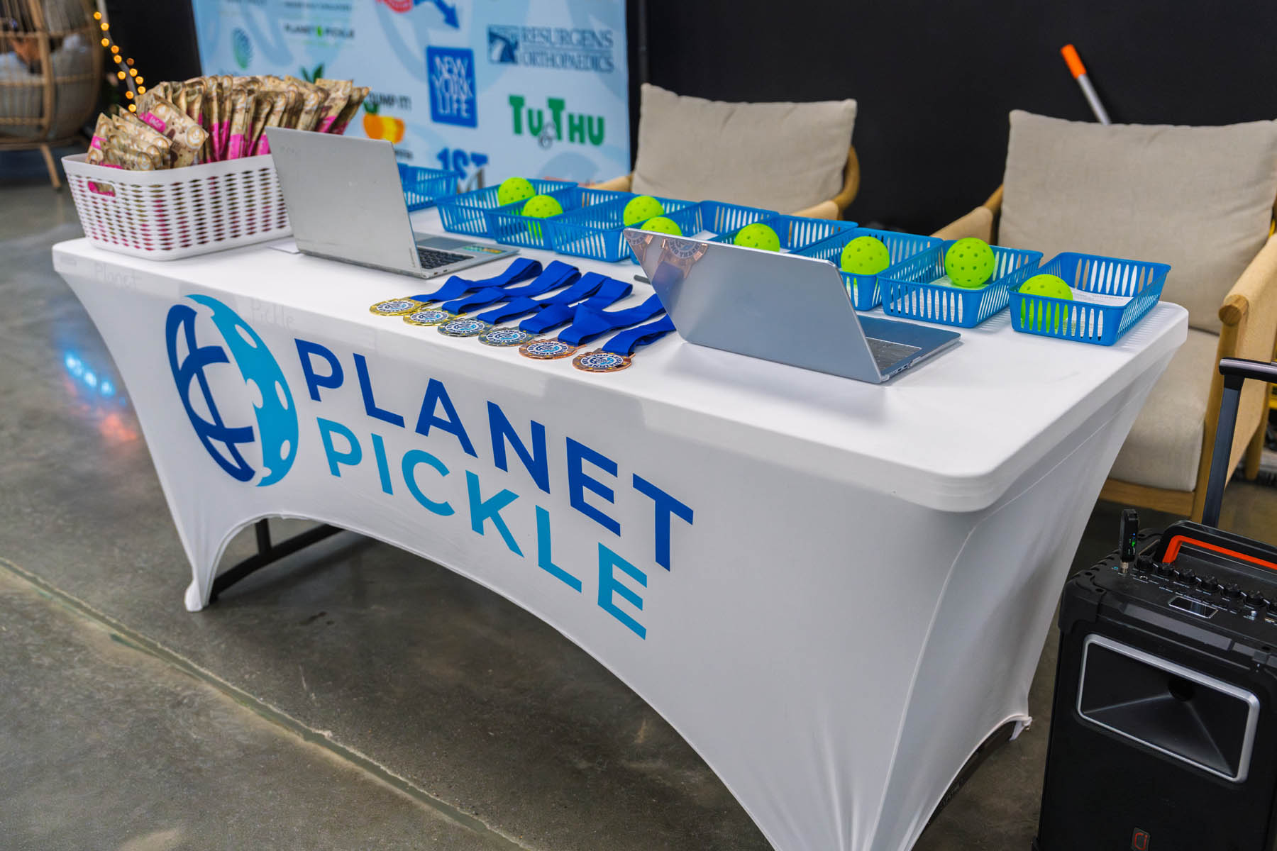 A table with a "Planet Pickle" cover displays medals, laptops, baskets of pickleballs, and a basket of paddles at an indoor event.