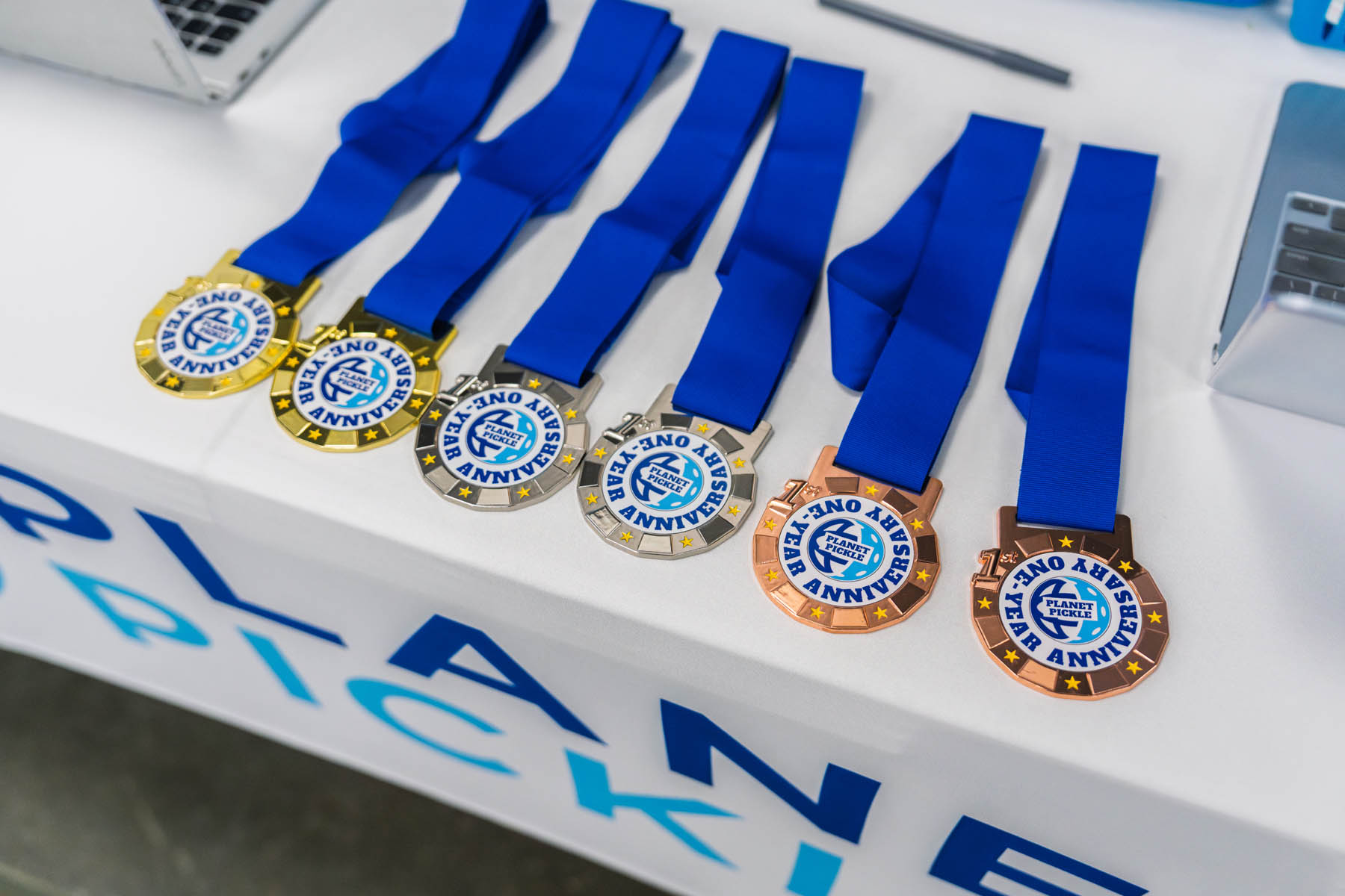 Six medals with blue ribbons, labeled "Plano Annual Pickleball," are displayed on a table, including gold, silver, and bronze varieties.