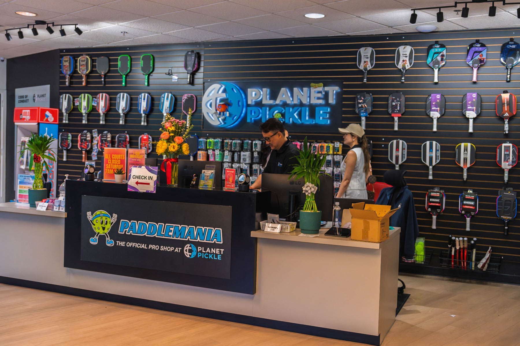 Two people stand behind the counter at a pro shop called Planet Pickle, with pickleball paddles and accessories displayed on the wall behind them.