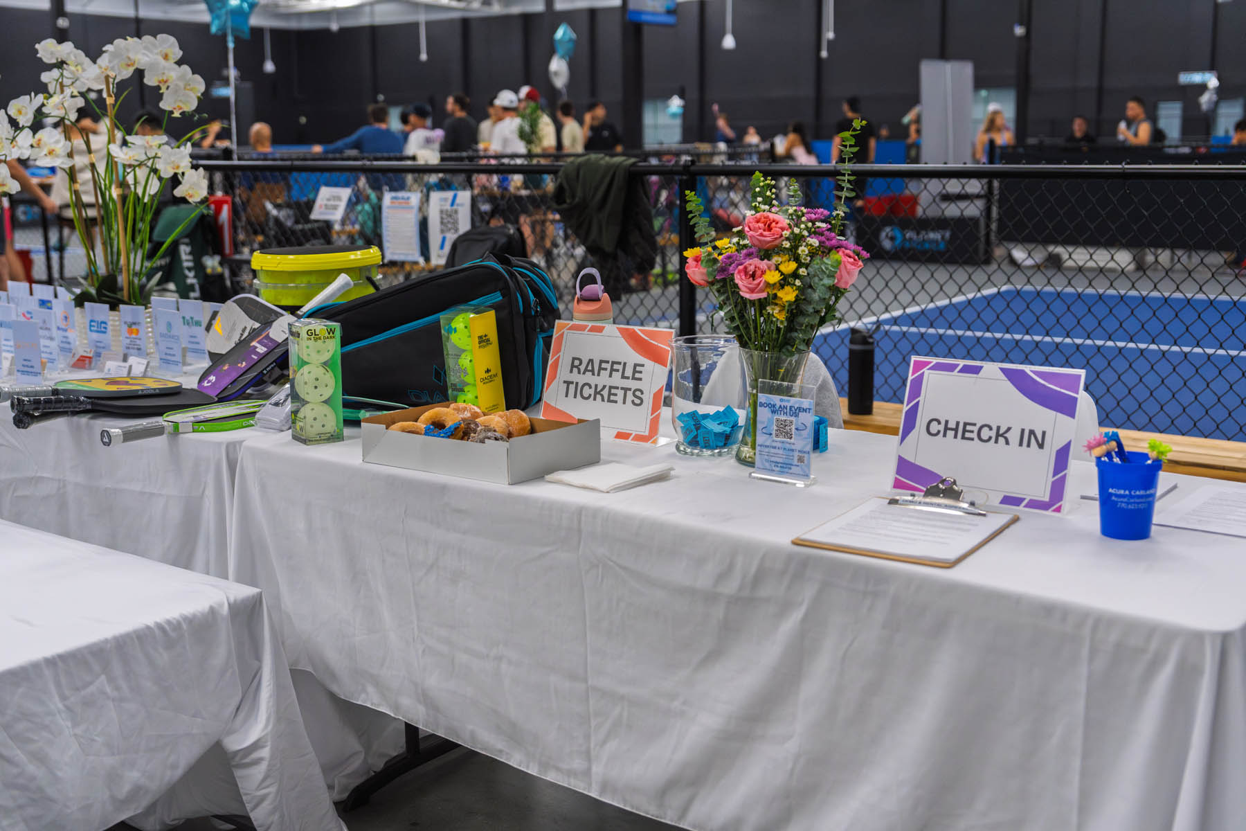 A table with raffle tickets, sign-in sheet, snacks, flowers, and prizes is set up at an indoor event with people and a tennis court in the background.