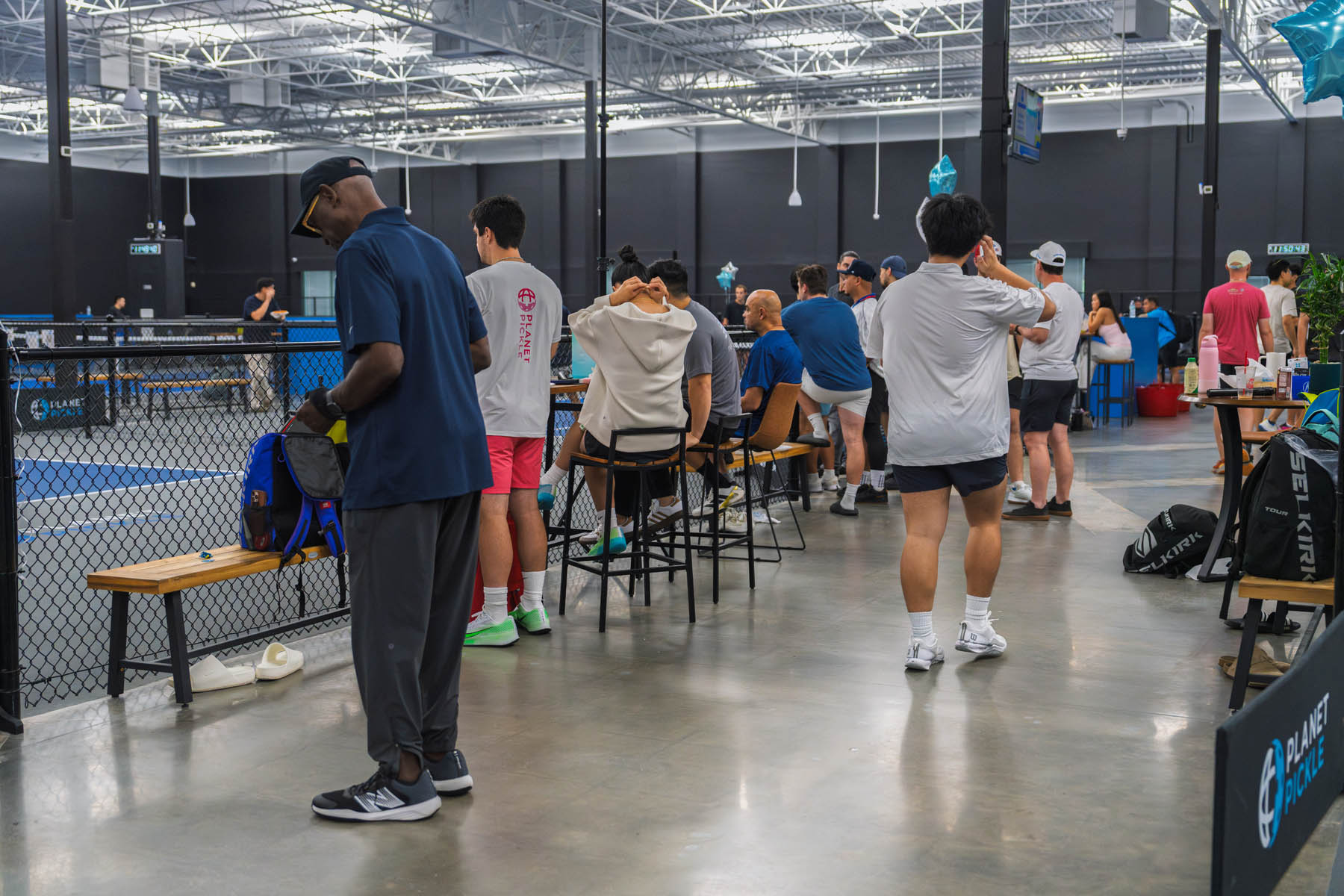 People prepare for a game inside an indoor sports facility, with some sitting on benches and others standing near a fenced pickleball court.