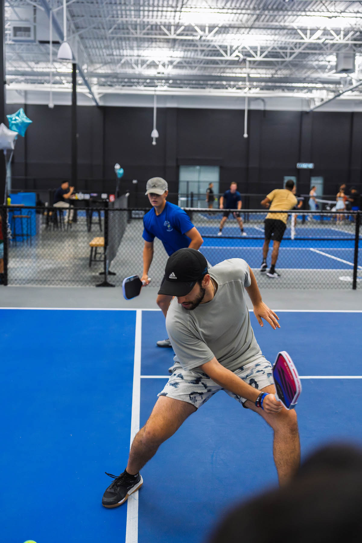 Two men play a game of pickleball indoors, with the player in front preparing to hit the ball while others play on adjacent courts in the background.