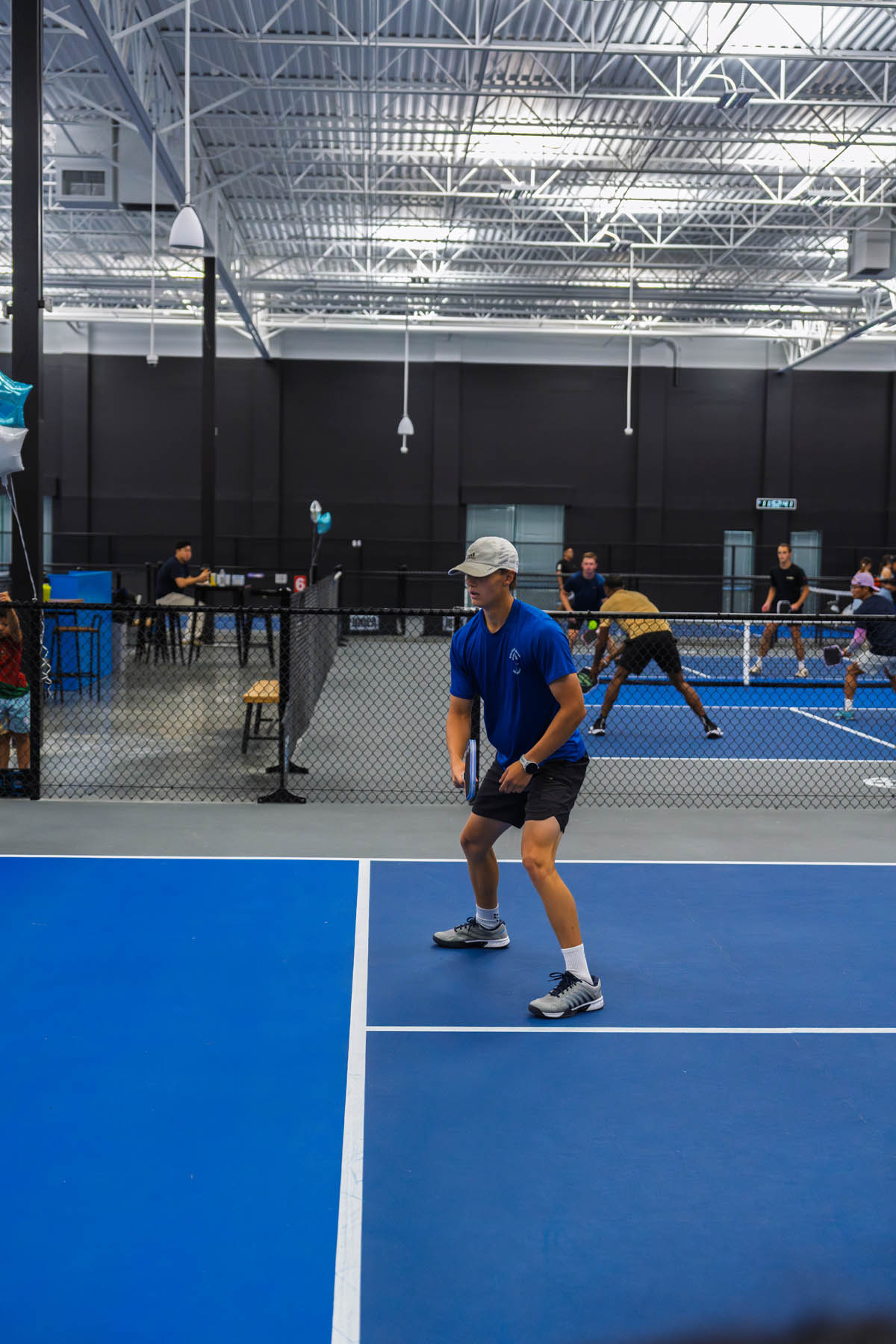 A man in athletic wear stands ready on a blue indoor court, holding a paddle, preparing for a game of pickleball. Other players and spectators are visible in the background.