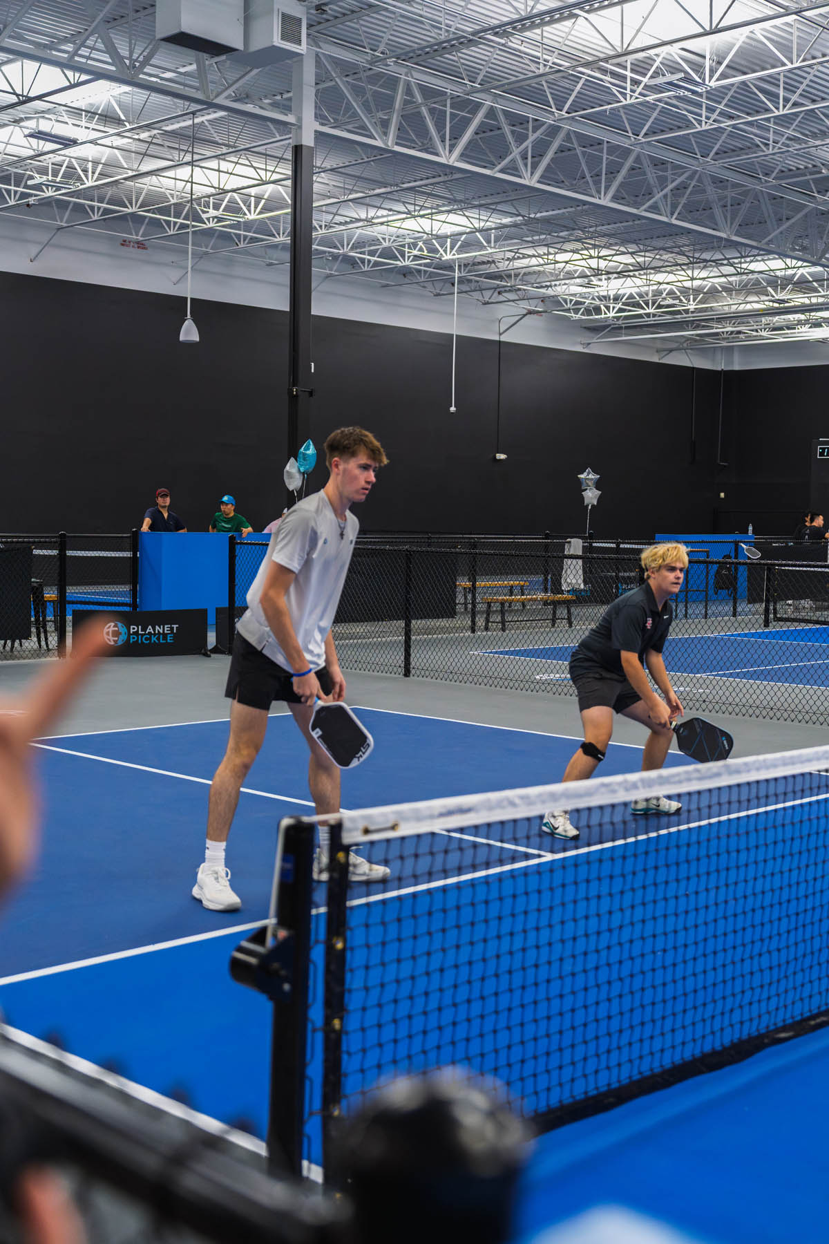 Two people stand ready with paddles on an indoor pickleball court, preparing to receive a serve during a doubles match.
