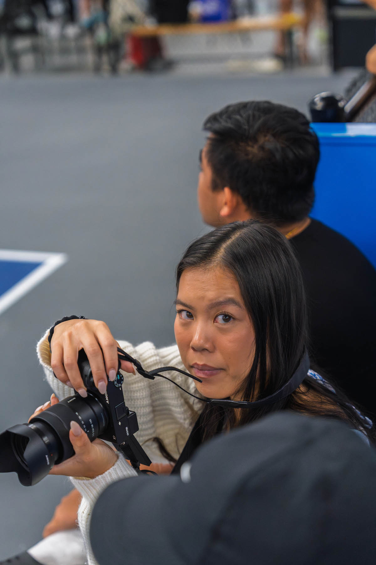 A woman holding a camera sits indoors, looking towards the lens, with two other people nearby.