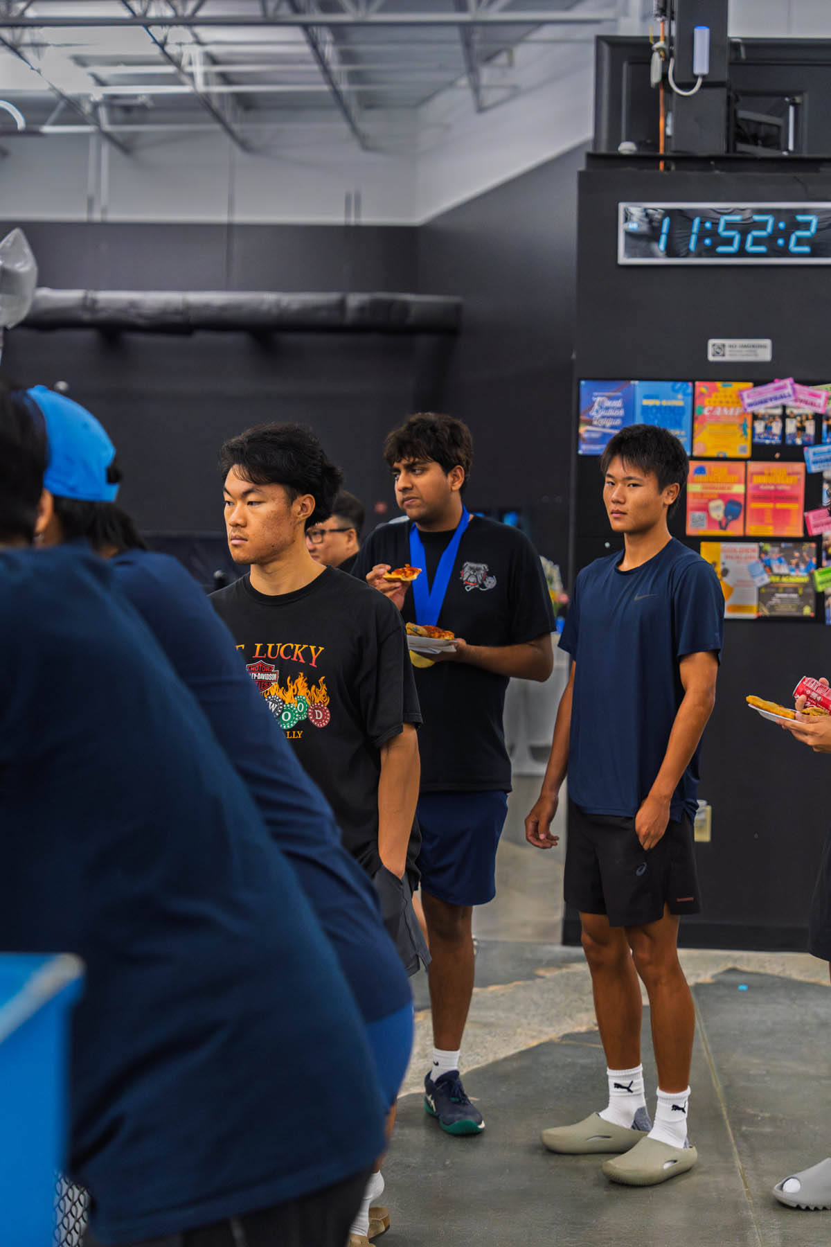 Several young men stand in line indoors, some holding plates of food. A digital clock on the wall shows 11:52; colorful flyers are posted nearby.