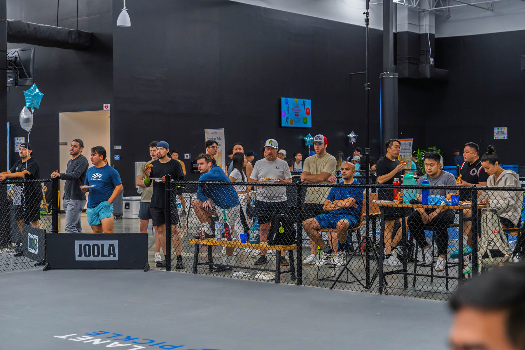 A group of people watch a pickleball game from behind a black fence inside an indoor sports facility, with tables, chairs, and sports equipment visible.