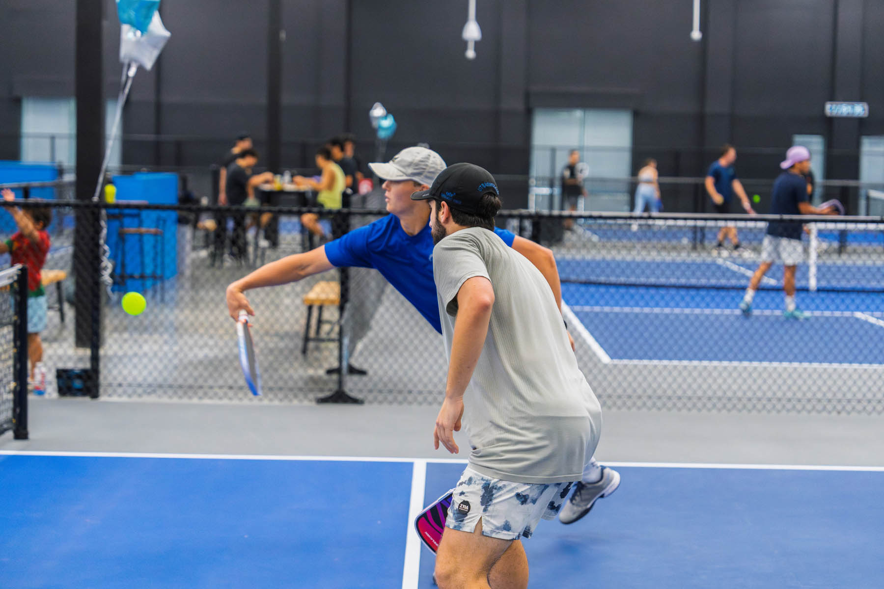 Two men play a doubles paddle tennis match indoors, with other players and courts visible in the background.