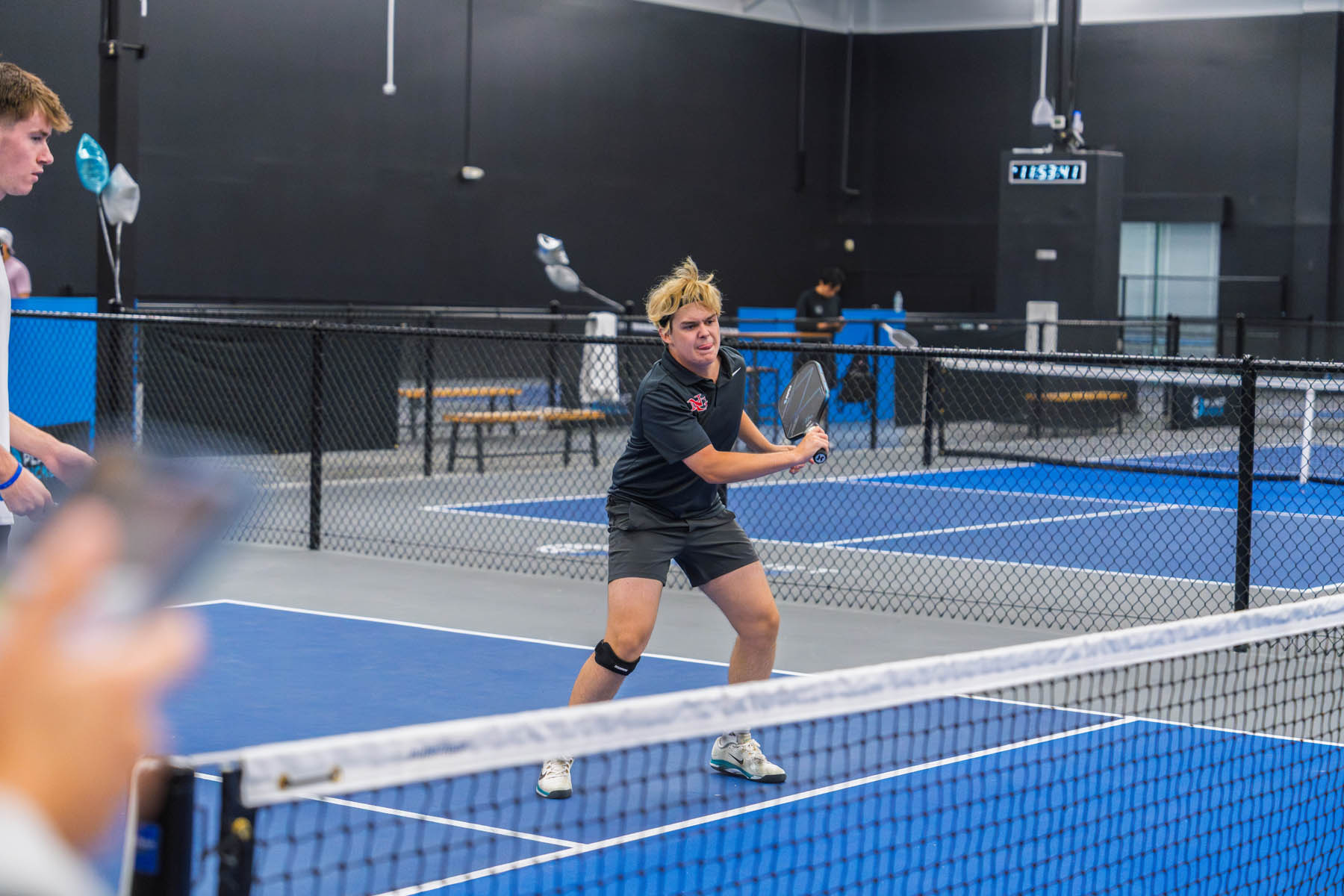A person prepares to hit a ball with a paddle on an indoor pickleball court, while another player stands nearby.