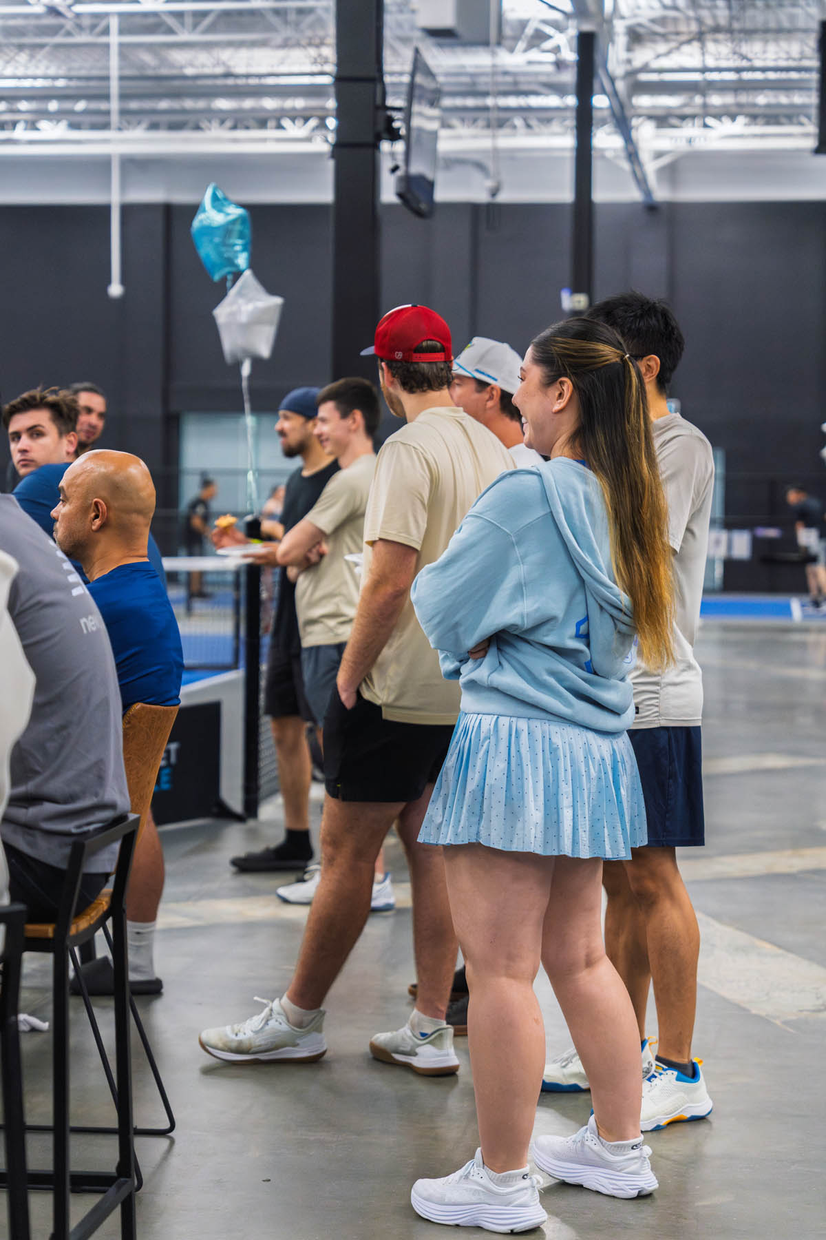 A group of people stand indoors near a table, some wearing athletic clothing, with blue and silver balloons visible in the background.