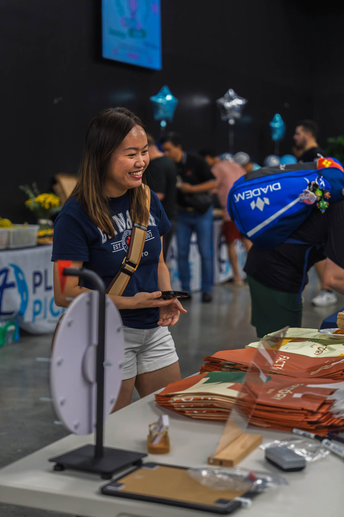 A woman stands at a table with promotional tote bags and a spinning prize wheel at an indoor event with other people in the background.
