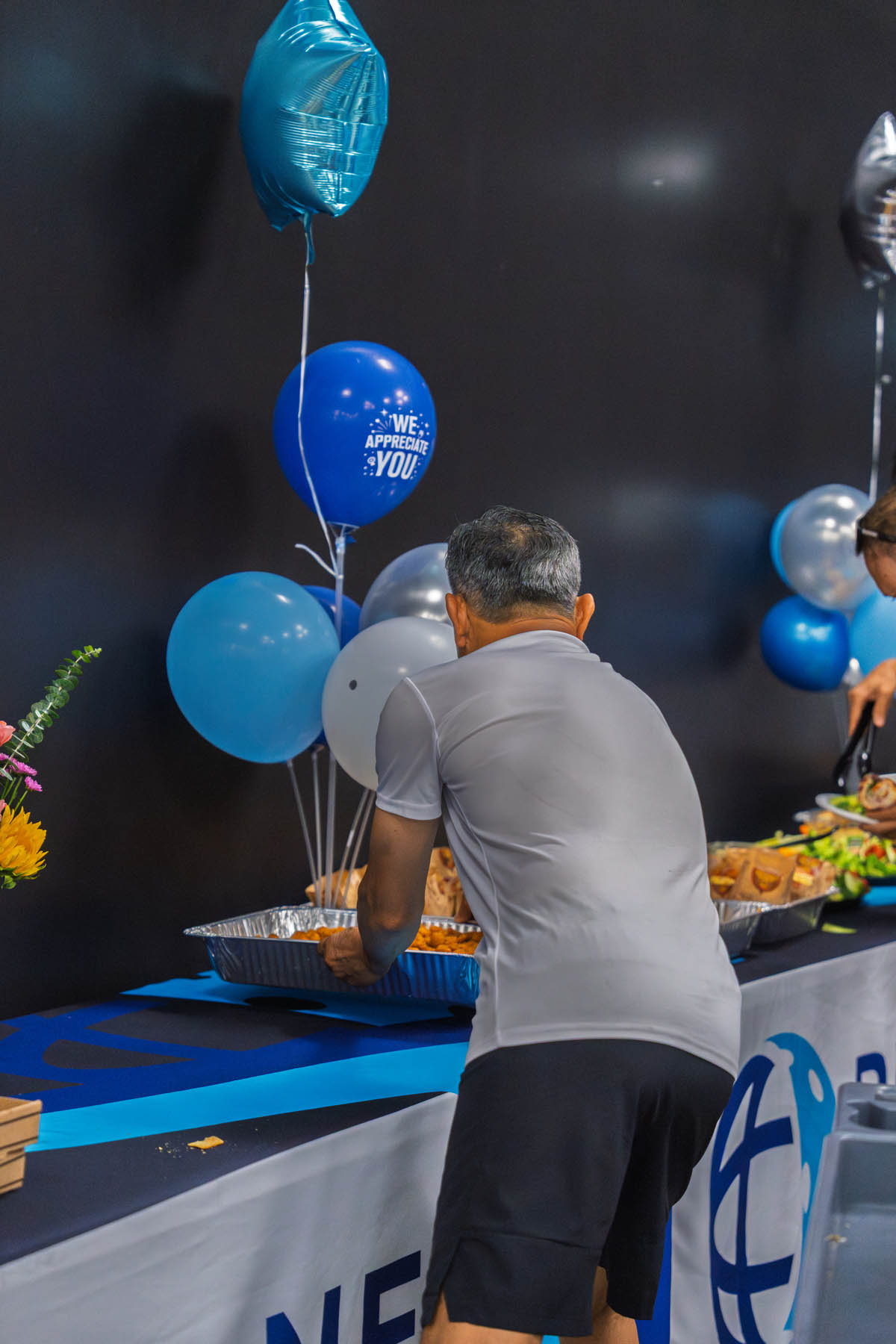 A man in athletic clothing serves food from a buffet table decorated with blue and silver balloons at an indoor event.