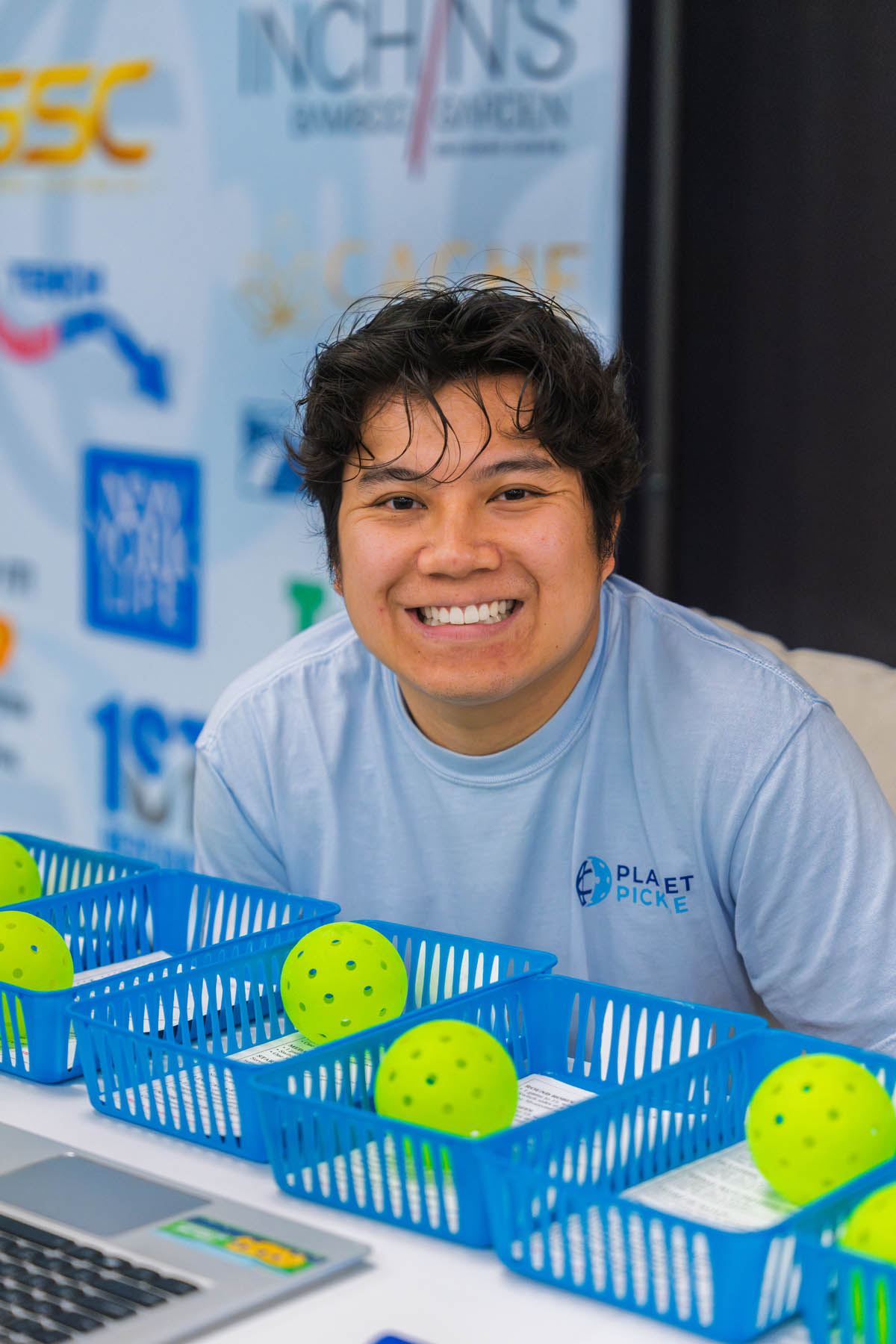 A person in a light blue "Planet Pickle" shirt sits behind a table with blue baskets containing yellow perforated balls, smiling at the camera.
