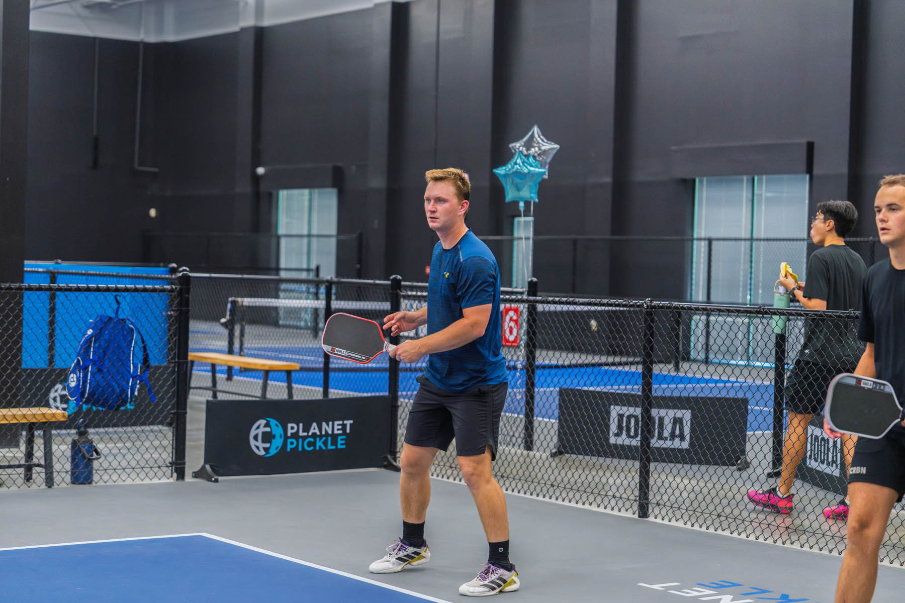 Two men stand ready with paddles on an indoor pickleball court, preparing for play. A balloon and benches are visible in the background.