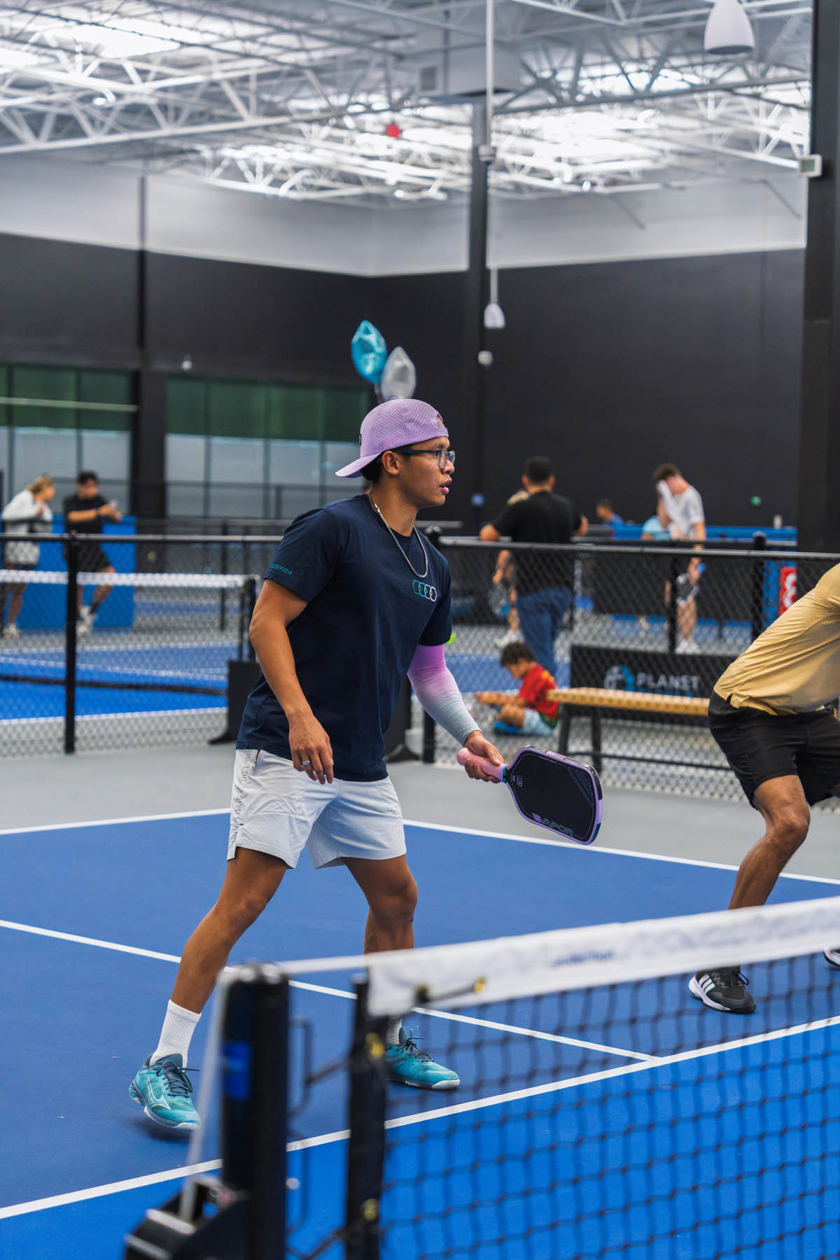 Two people play pickleball indoors on a blue court, with others visible in the background. One player is ready with a paddle, wearing a cap, glasses, and a black shirt.