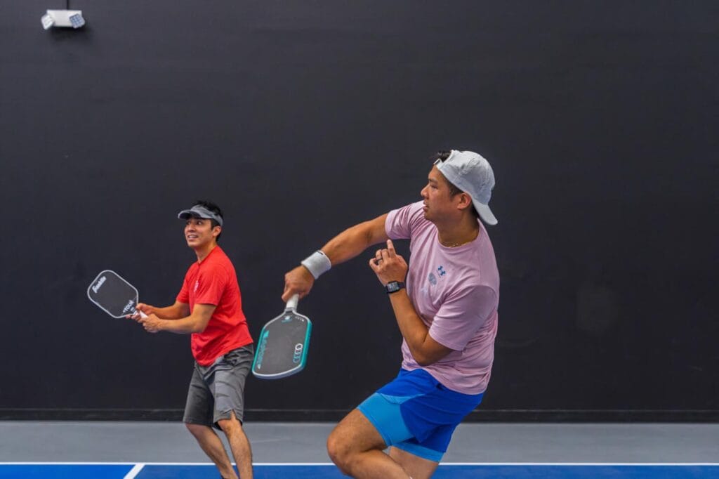 Two men play pickleball indoors; one in a red shirt stands ready while the other in a pink shirt swings his paddle, both on a blue court with a dark wall background.