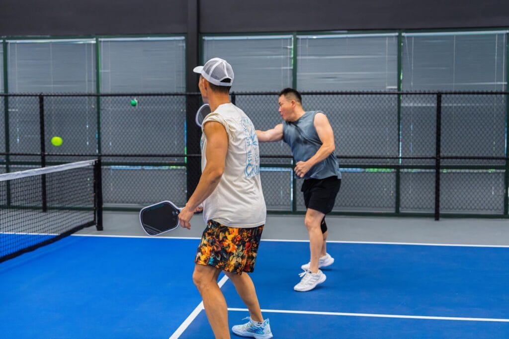 Two men playing pickleball indoors; one is about to hit the ball while the other stands ready near the net.