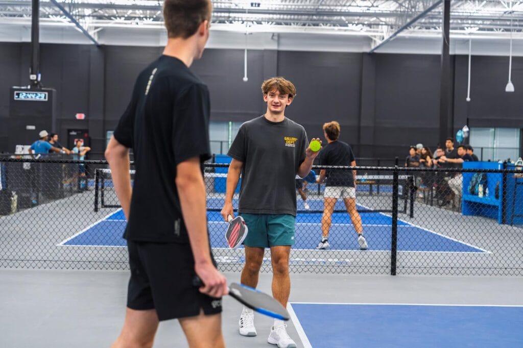 Team Building & Fun: How to Host Your Corporate Pickleball Events 6 Two young men stand on an indoor pickleball court holding paddles; one holds a ball and smiles while others play in the background.