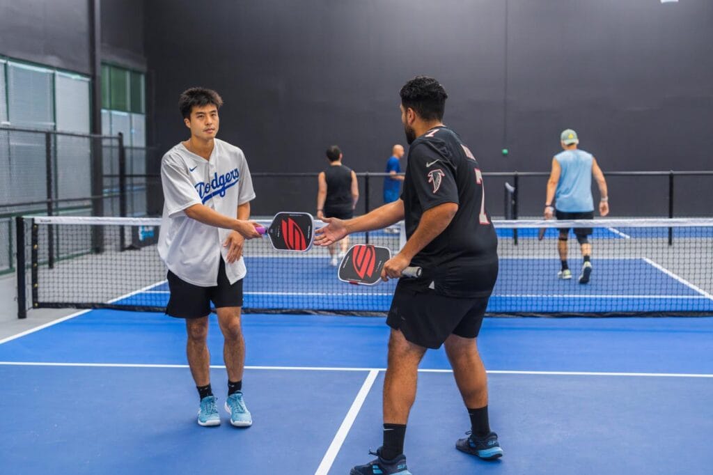 Two men on an indoor pickleball court tap paddles at the net while two other pairs play in the background.