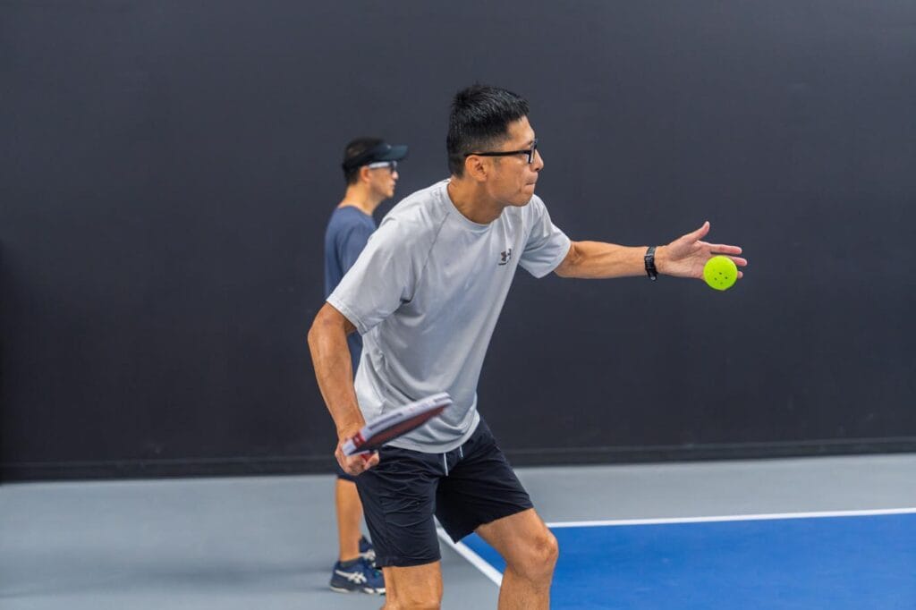 Team Building & Fun: How to Host Your Corporate Pickleball Events 7 A man in athletic wear prepares to hit a pickleball with a paddle on an indoor court, while another person stands in the background.