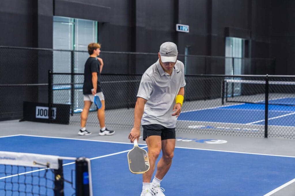 Two men playing pickleball indoors, one preparing to hit the ball with his paddle while the other stands near the back of the court.