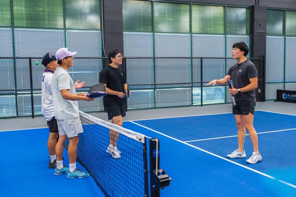 Team Building & Fun: How to Host Your Corporate Pickleball Events 8 Four men stand on opposite sides of a tennis court net, with one gesturing as if explaining, while the others listen and hold paddles.