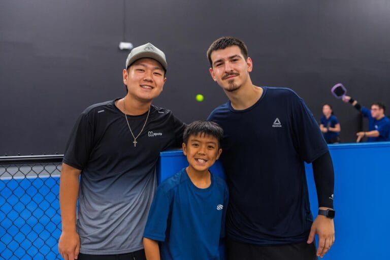 Three people, two adults and one child, stand together and smile at an indoor sports facility with others playing a game in the background.