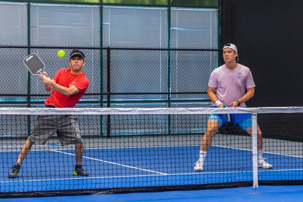 Team Building & Fun: How to Host Your Corporate Pickleball Events 4 Two men play doubles pickleball indoors; one in a red shirt prepares to hit the ball while the other in a pink shirt stands ready beside him.