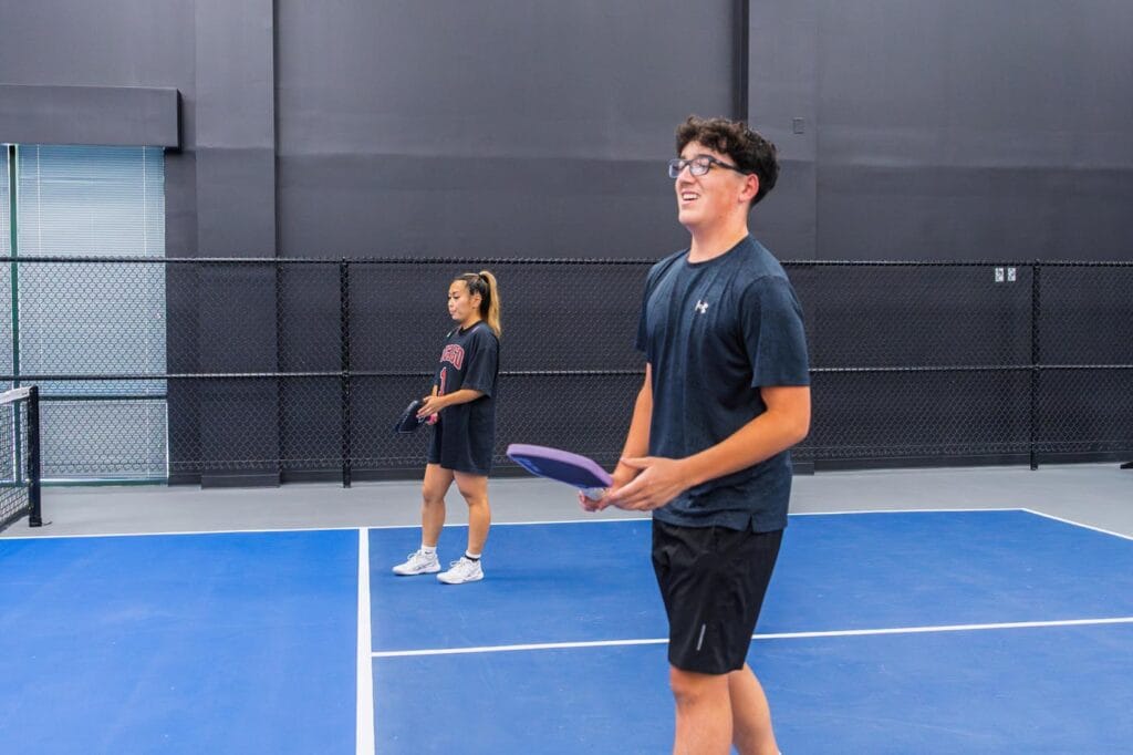 Two people stand on an indoor pickleball court holding paddles, with a net and black fence in the background.